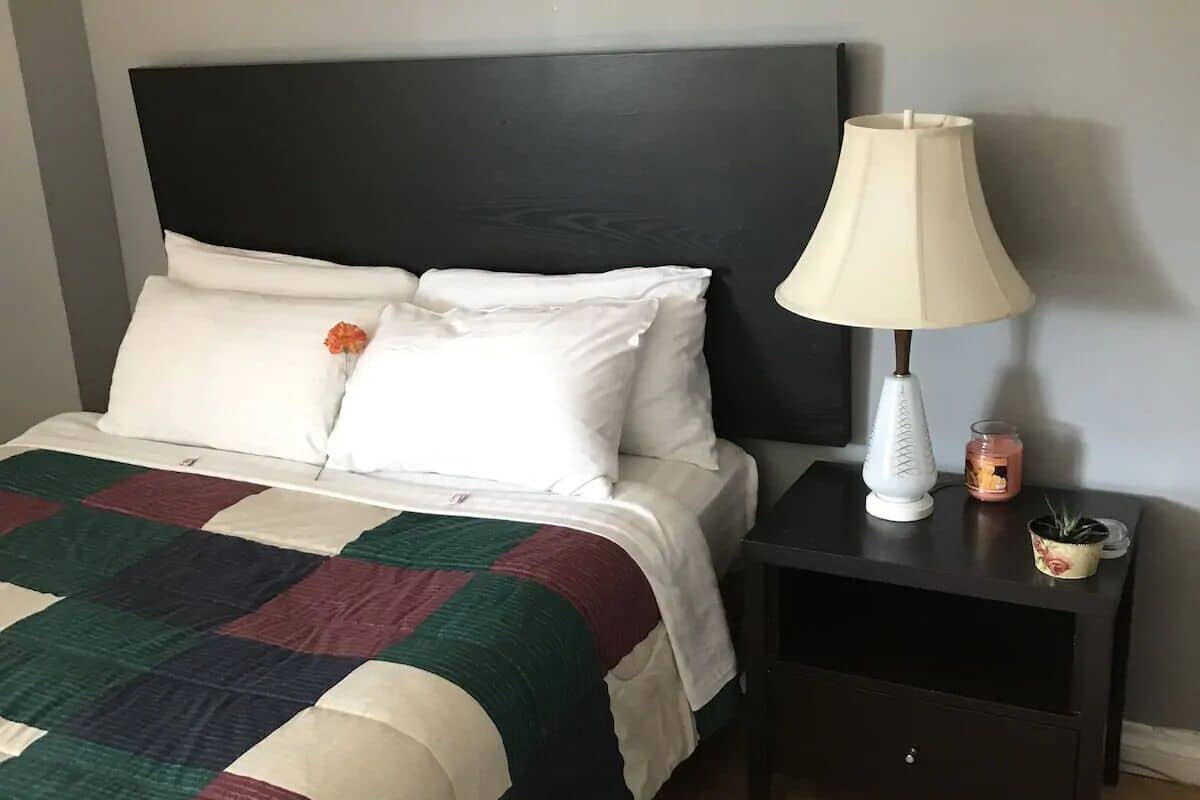 Una cama con colcha, una mesita de noche con lámpara, un florero y una planta pequeña.