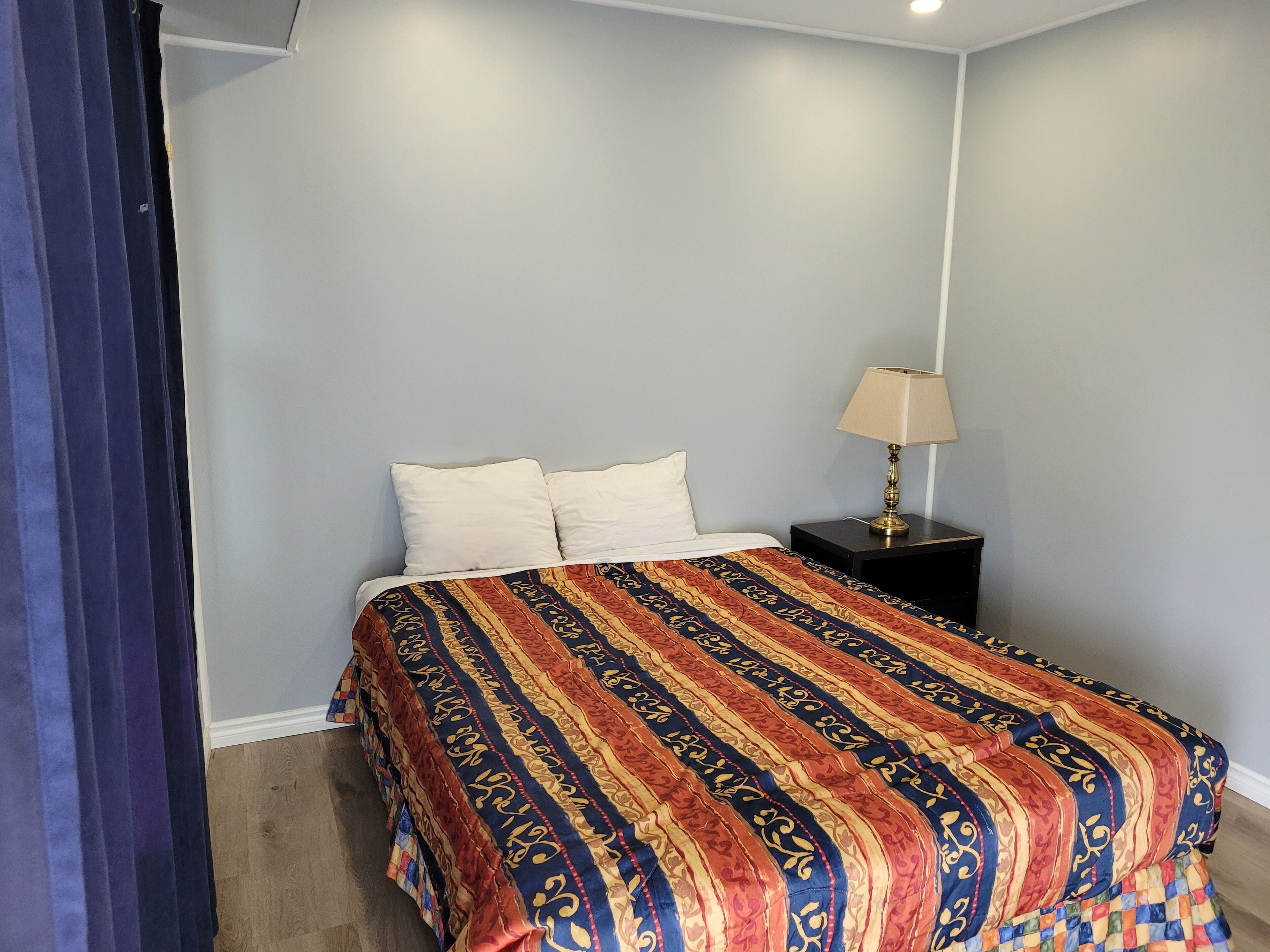 Un dormitorio con una cama, dos almohadas, una colcha estampada, una mesita de noche con una lámpara y una cortina oscura.