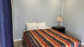 Un dormitorio con una cama, dos almohadas, una colcha estampada, una mesita de noche con una lámpara y una cortina oscura.