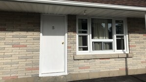 Ein einstöckiges Backsteinhäuschen mit einer weißen Tür, auf der die Zahl 5 steht, einem mit Vorhängen versehenen Fenster und einer kleinen Veranda.