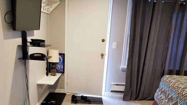 Ein Hotelzimmer mit Flachbildfernseher an der Wand, einem Schreibtisch mit Lampe, einem Bett mit gemusterten Bettwaren und einer Tür zu einem weiteren Raum.