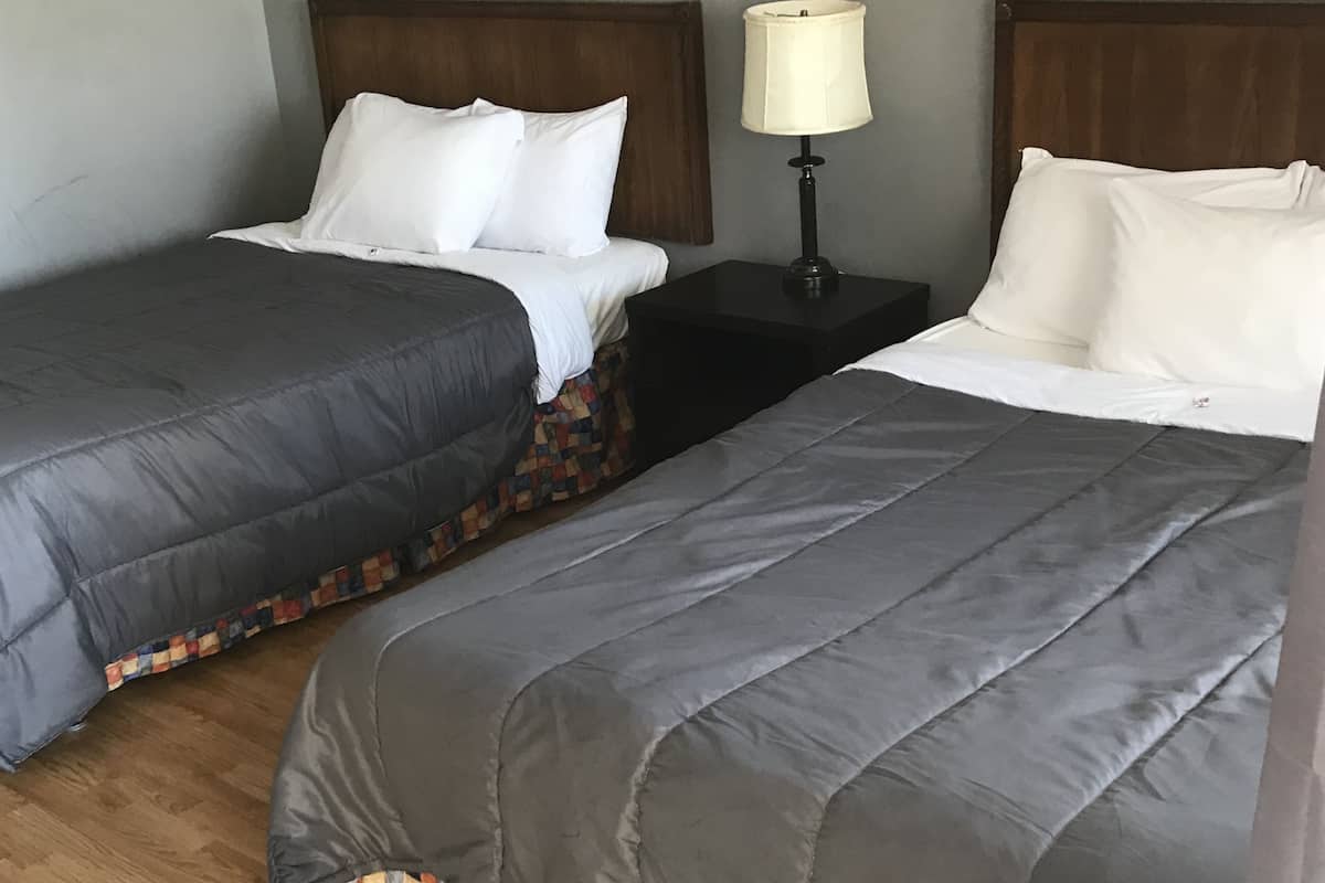 Dos camas individuales con ropa de cama gris, una mesita de noche pequeña con una lámpara y un piso de madera.