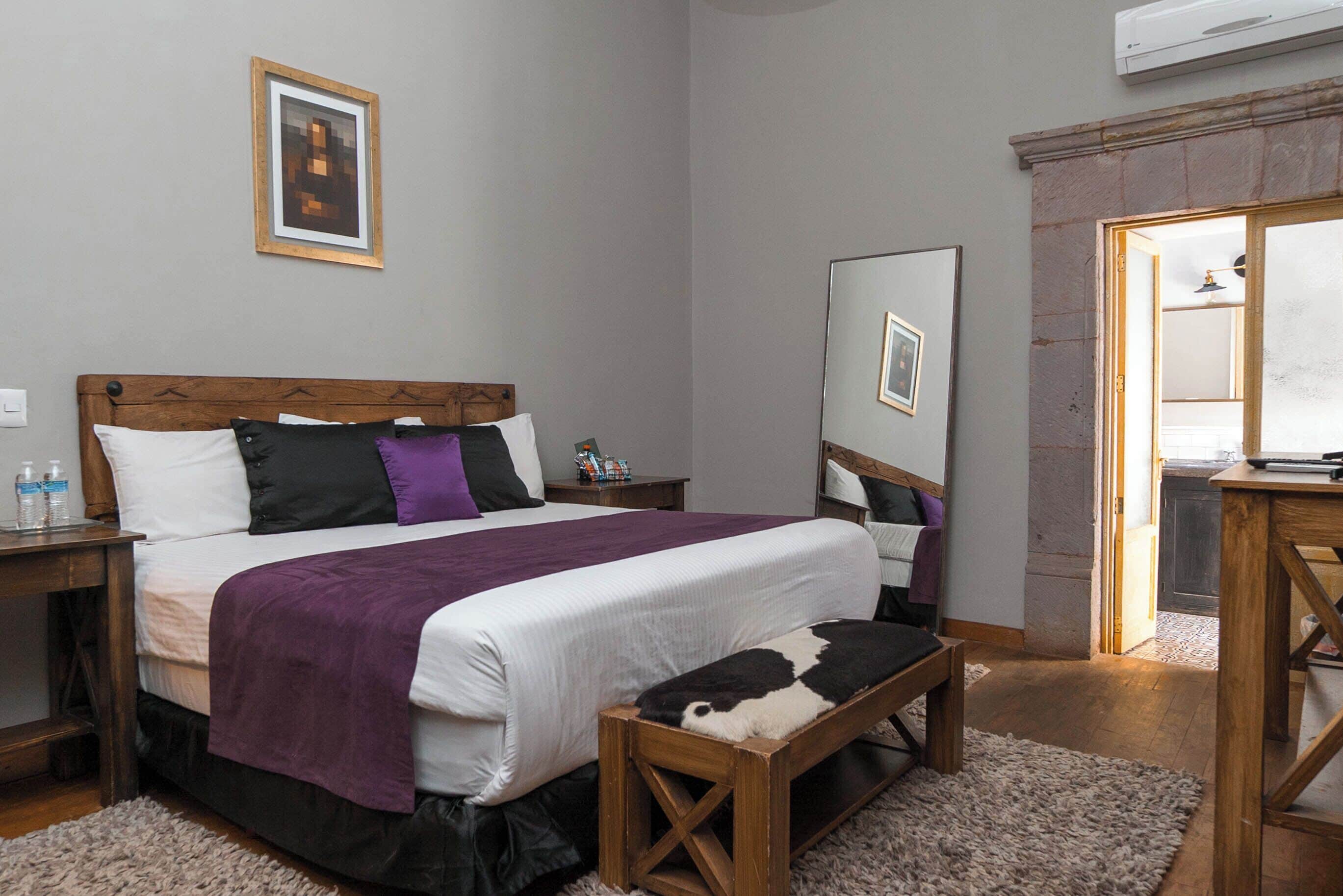Un dormitorio con cabecera de madera, una cama grande con ropa de cama blanca y morada, un banco de madera, un espejo y una mesita de noche con artículos de aseo.
