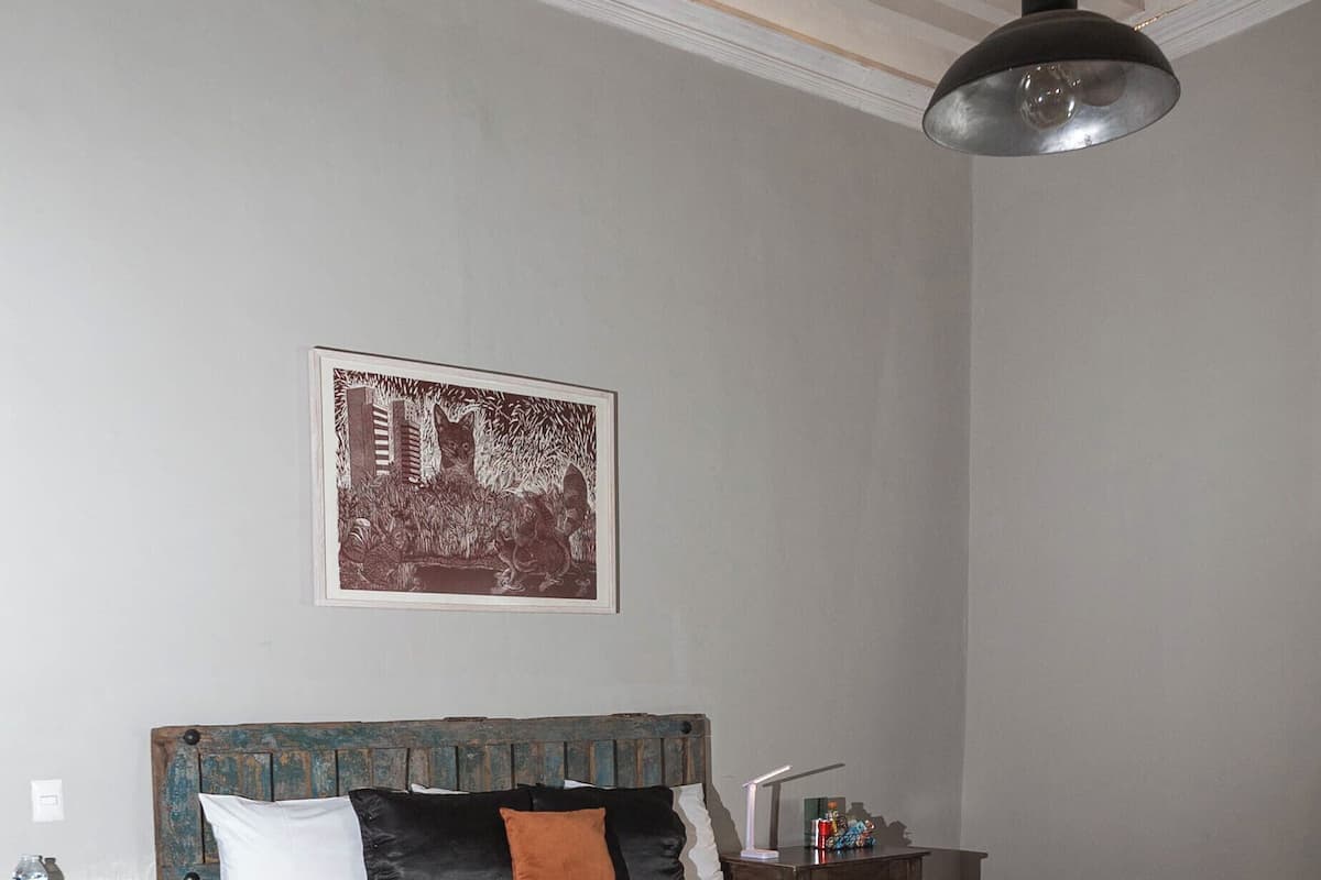 Un dormitorio ordenado con una cama grande, un banco de madera, una mesita de noche, una lámpara de pared y un cuadro enmarcado sobre la cama.