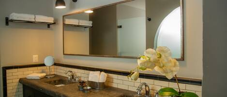 A bathroom with two sinks, a large mirror, a towel rack, a small round mirror, and a potted orchid.