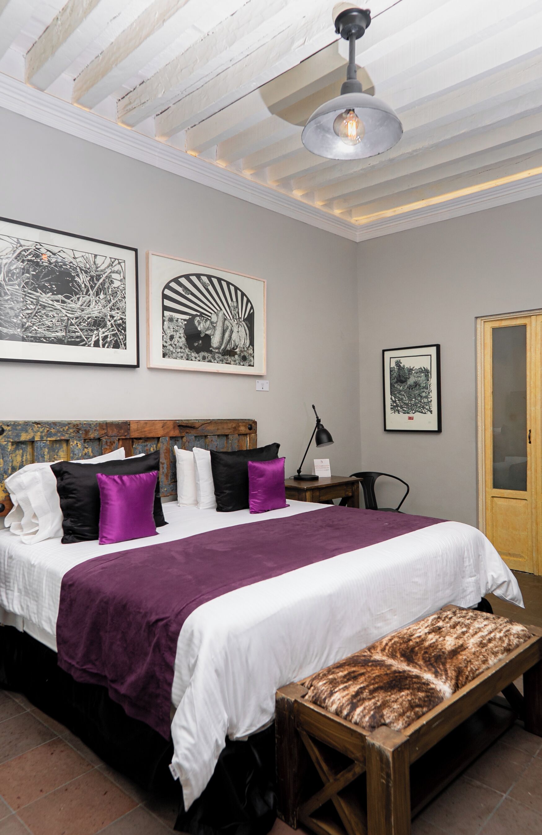 Un dormitorio con una cama, un banco, un escritorio y obras de arte en las paredes.