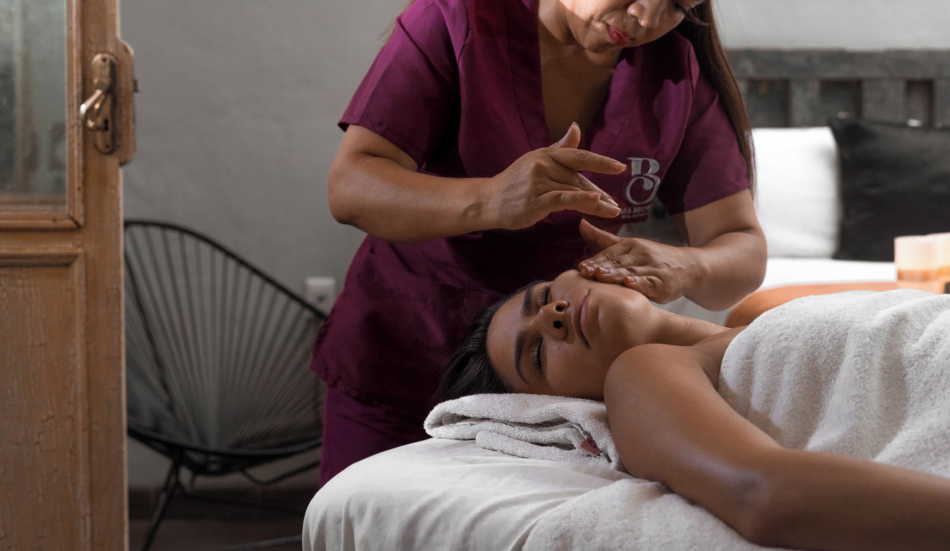 A person receiving a facial treatment in a room with a bed, a chair, and a door.