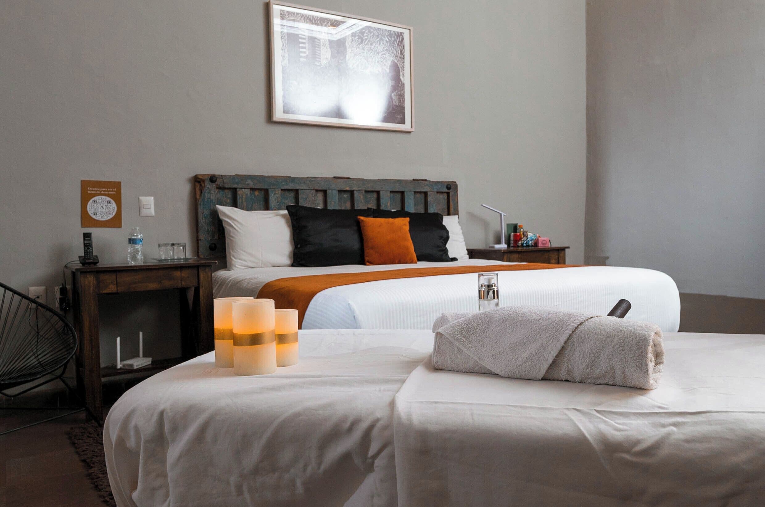 Habitación de hotel con una cama grande, cabecera de madera, ropa de cama blanca, una almohada blanca, una almohada naranja, una toalla blanca, una mesita de noche con lámpara, un cuadro enmarcado en la pared y una silla.