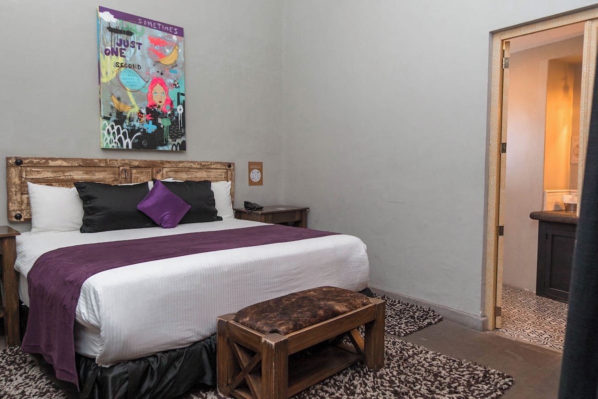 Un dormitorio con cabecera de madera, cama con ropa de cama blanca, almohada morada, un banco y un cartel en la pared.