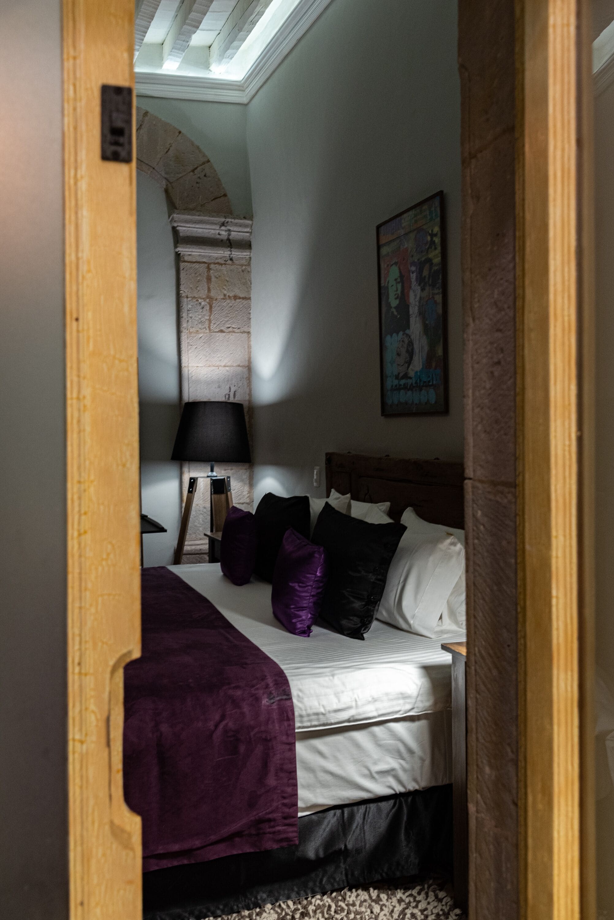 A bedroom with a bed, a lamp, a painting, and a stone wall.