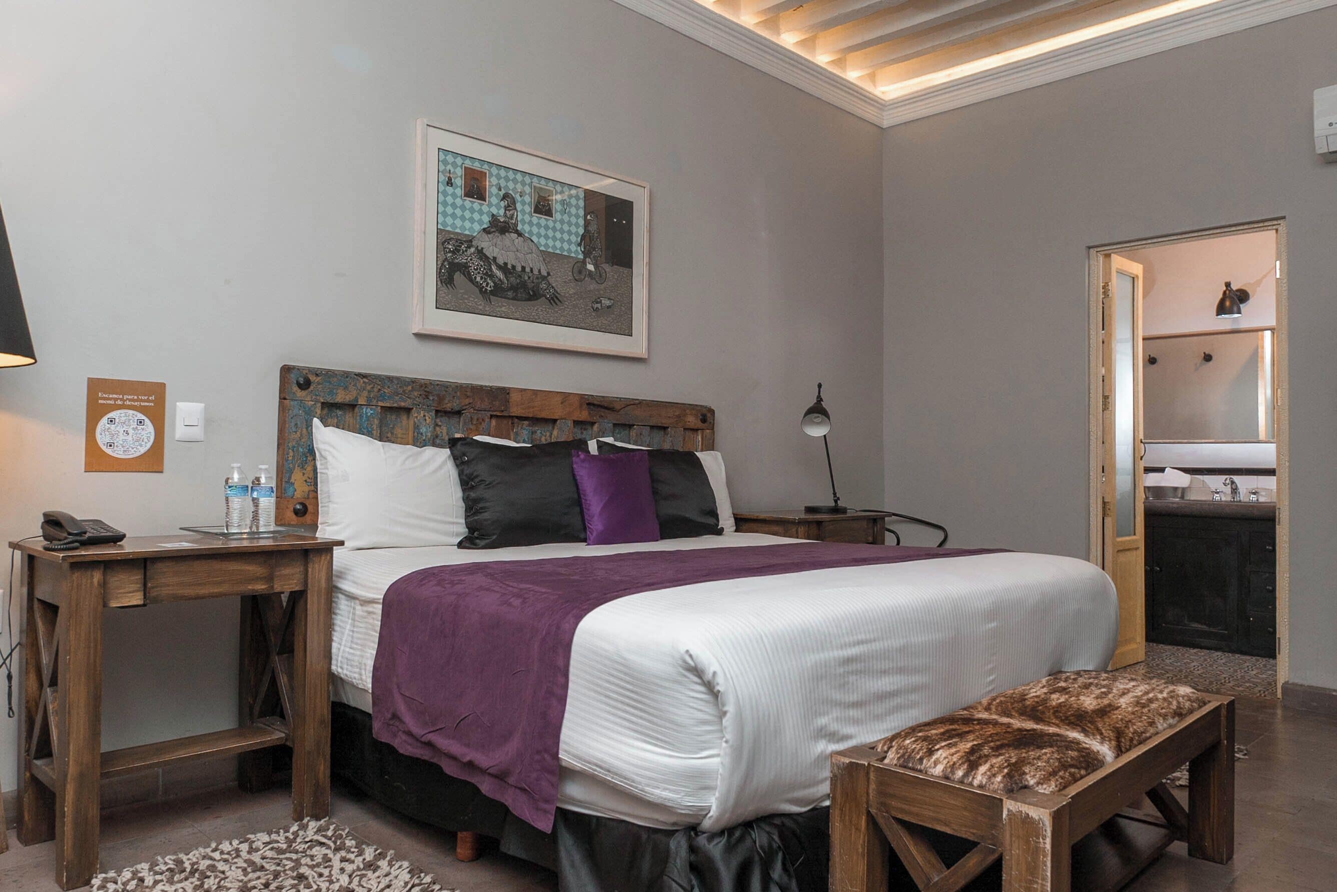 Un dormitorio con cama, mesitas de noche, un banco, un cuadro en la pared y una puerta que da al baño.
