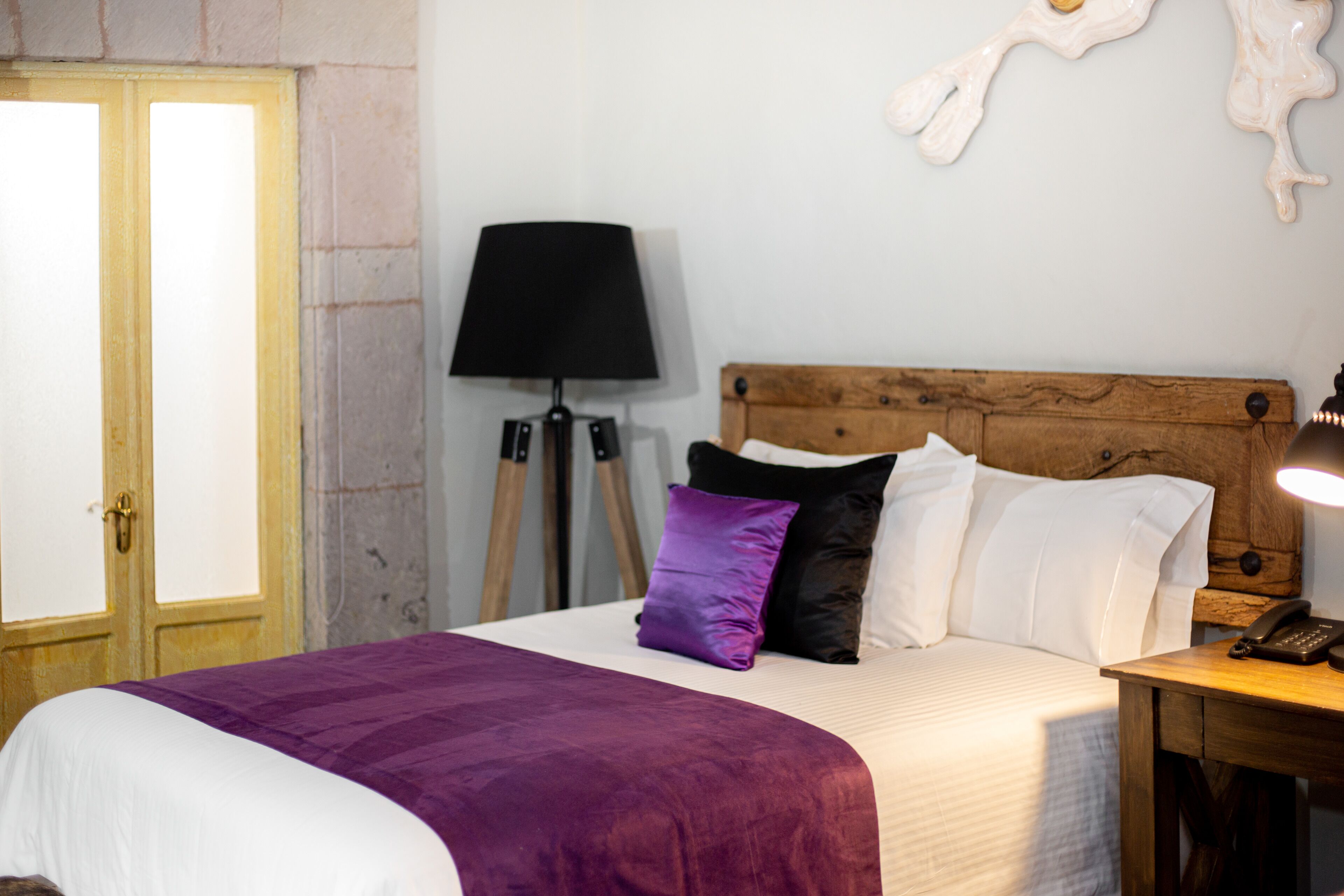 Un dormitorio con cabecera de madera, cama con ropa de cama blanca y morada, una lámpara y un teléfono en una mesita de noche.