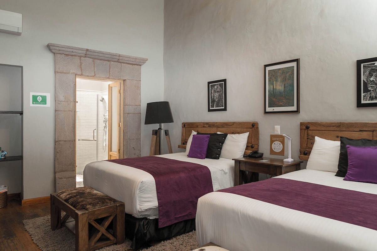 Habitación de hotel con dos camas, chimenea de piedra, banca de madera y obras de arte enmarcadas en las paredes.