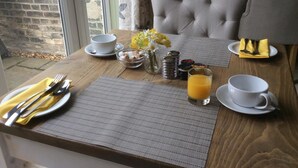 Ein Frühstückstisch mit Holzplatte, zwei grauen Sitzgelegenheiten, einer Vase mit gelben Blumen, einem Glas Orangensaft und ordentlich drapierten Frühstücksleckereien.