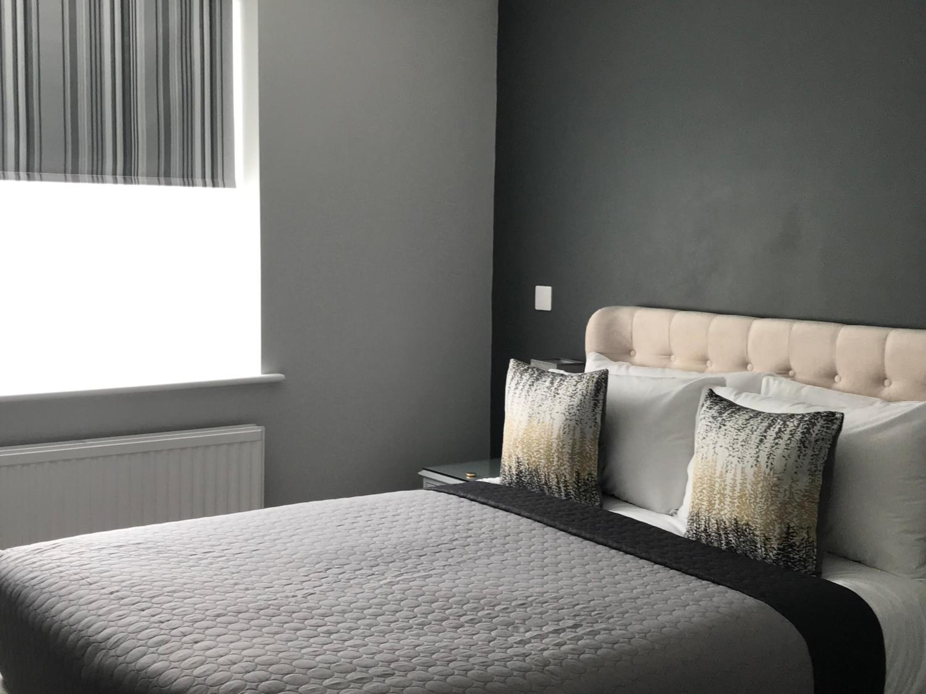 A bedroom with a bed, a window with blinds, a radiator, and a nightstand with a lamp.