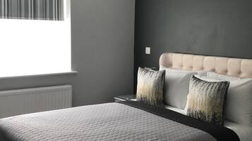A bedroom with a bed, a window with blinds, a radiator, and a nightstand with a lamp.