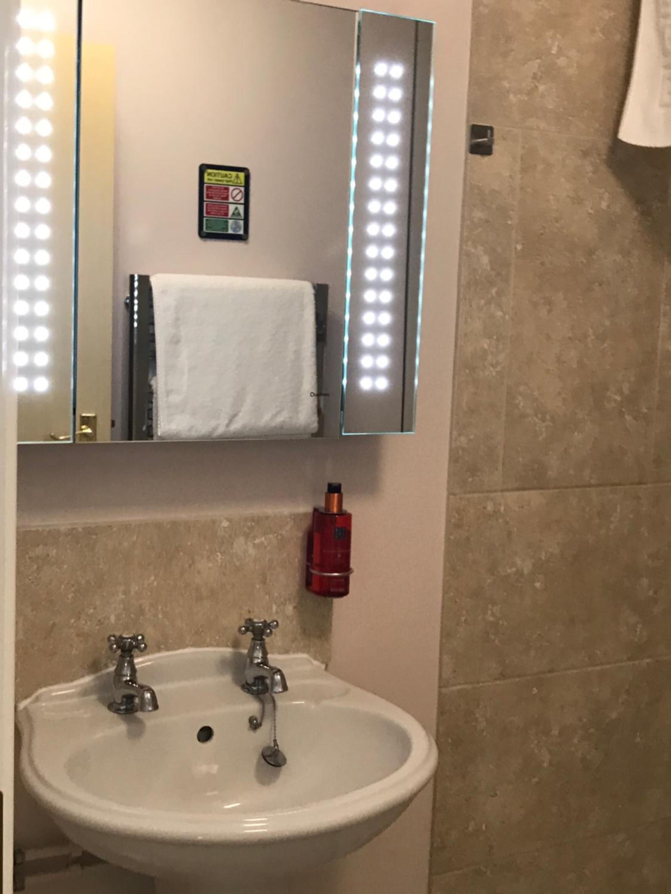 Uma casa de banho com uma pia branca, duas torneiras, um espelho iluminado e um porta-toalhas.