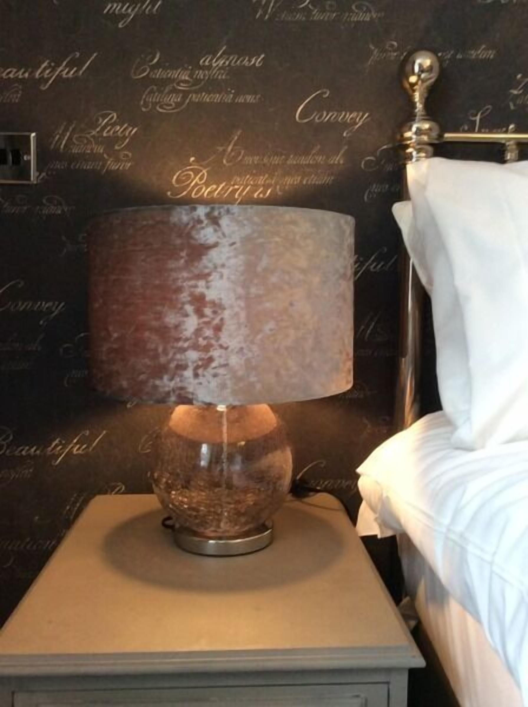 A bedside table with a vintage-style lamp, a white bed, and a wall with repeated text.