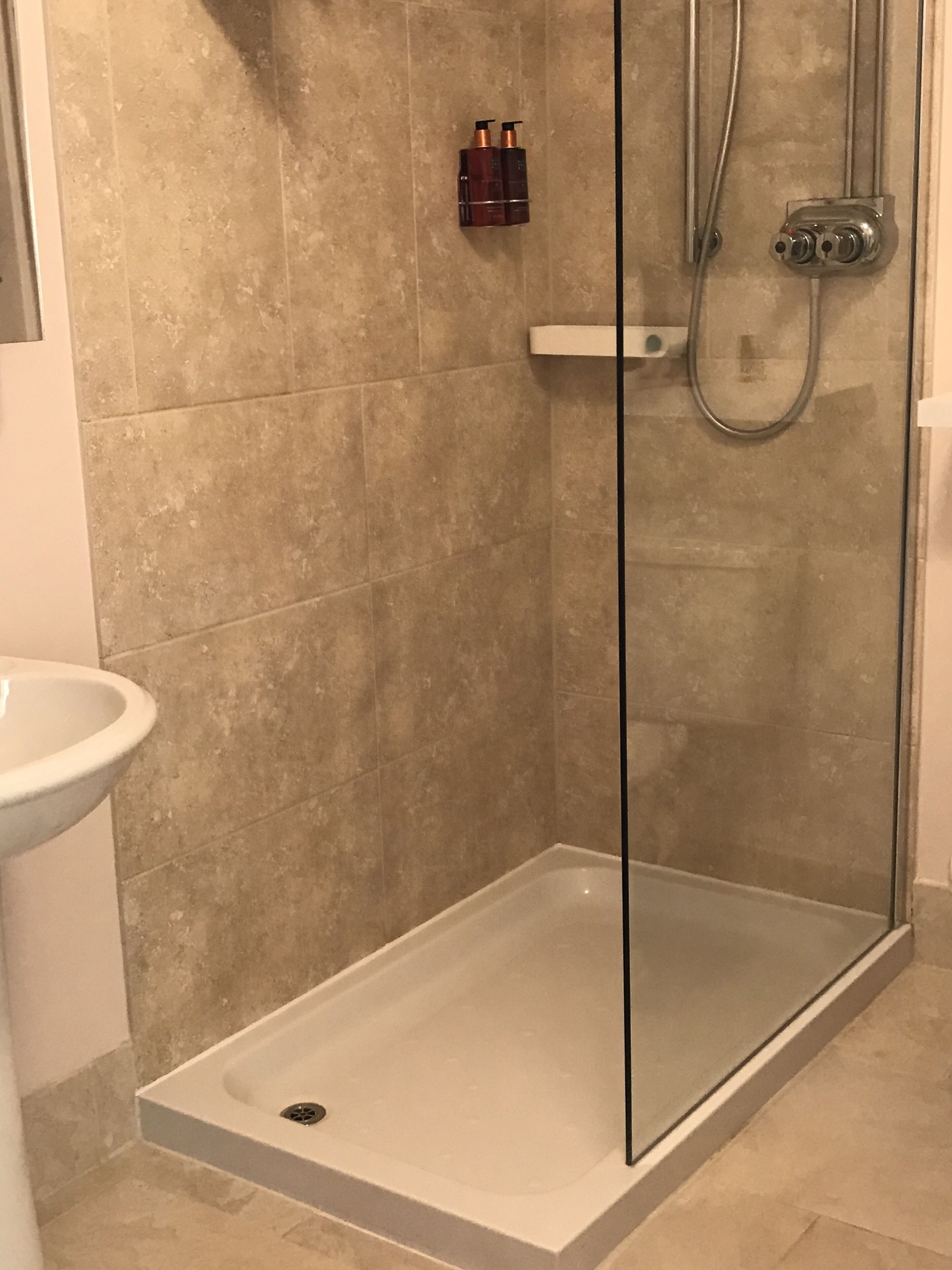 Een douche met een glazen deur, een witte wastafel en twee shampoo flessen aan de muur.
