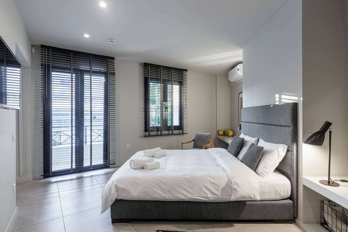 Une chambre à coucher moderne avec un grand lit, un bureau avec une lampe et une vue à travers les stores de la fenêtre.