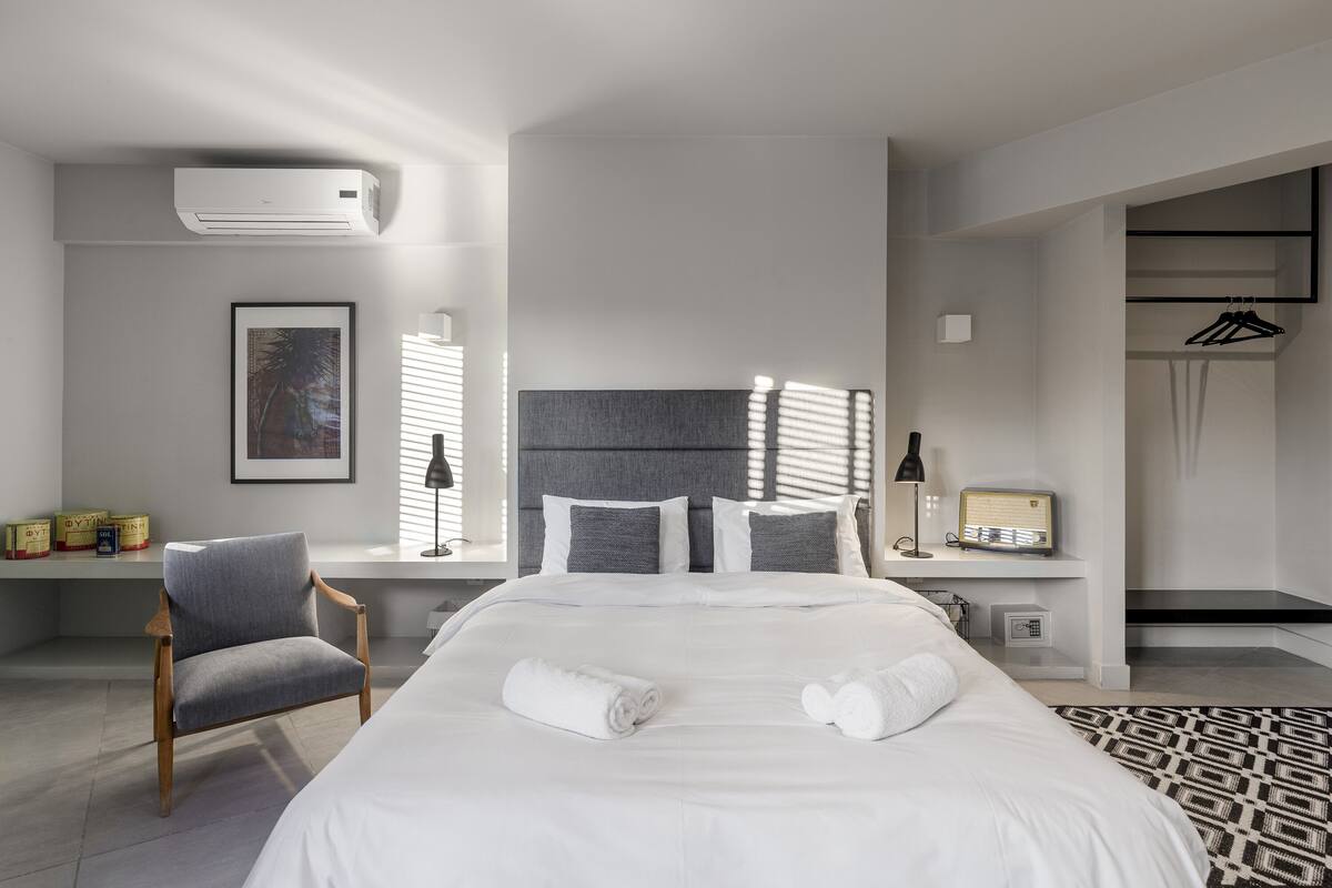 Une chambre d’hôtel moderne avec un grand lit, un fauteuil gris, une table de chevet avec une lampe et une petite radio.