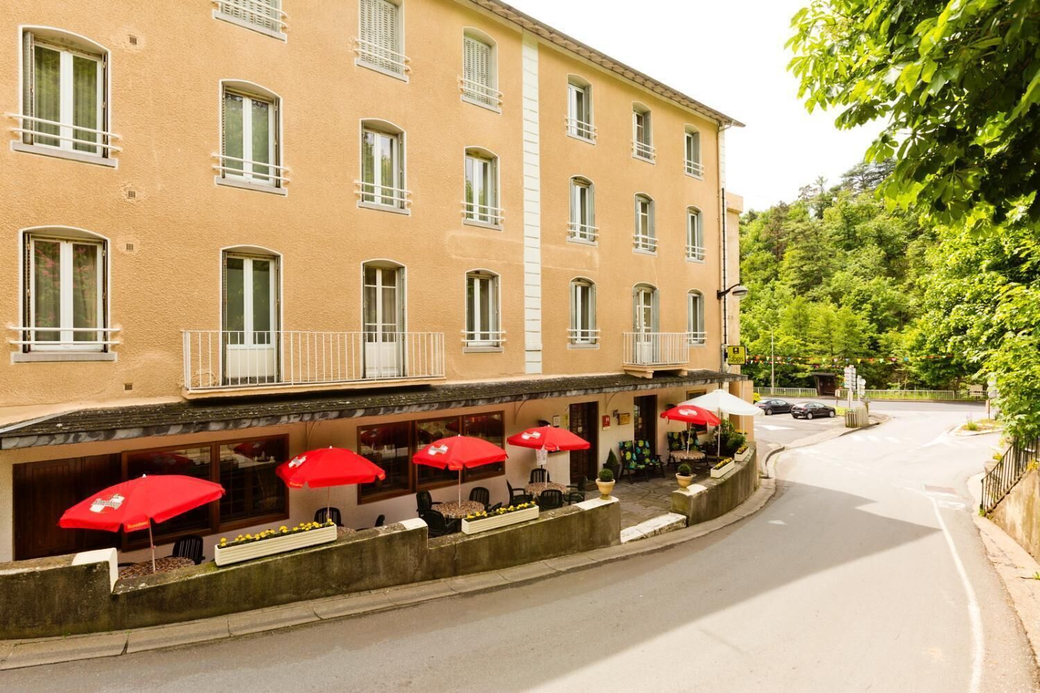 A building with multiple windows and balconies, outdoor seating with red umbrellas, and a street view with cars and greenery.