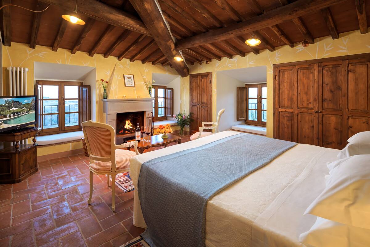 Un dormitorio amplio con una cama grande, vigas de madera en el techo, una chimenea y un televisor sobre un mueble de madera.