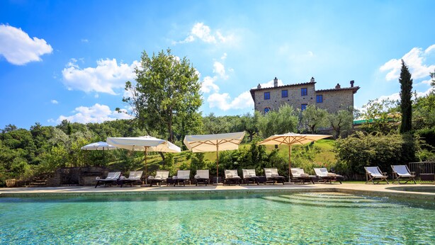 A pool area with lounge chairs and umbrellas, surrounded by greenery and a stone building in the background. - Le Lappe (Cavriglia)