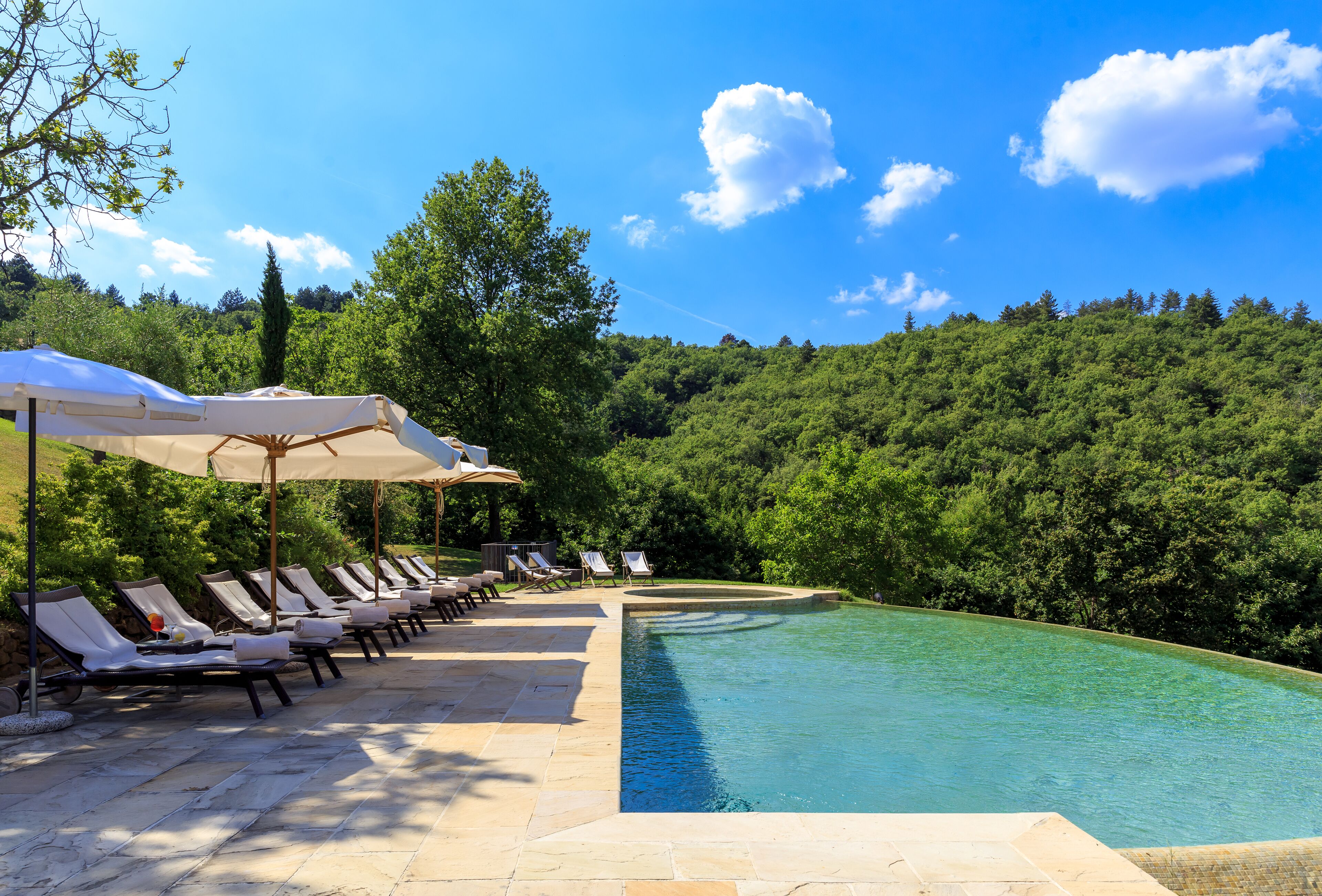Zona de piscina amb gandules i ombrel·les, envoltada de vegetació exuberant i un cel blau clar.