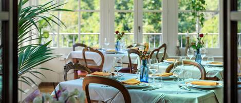 A dining area with tables set for a meal, large windows, and potted plants.