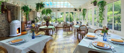 A conservatory with tables set for dining, a stone fireplace, and hanging plants.