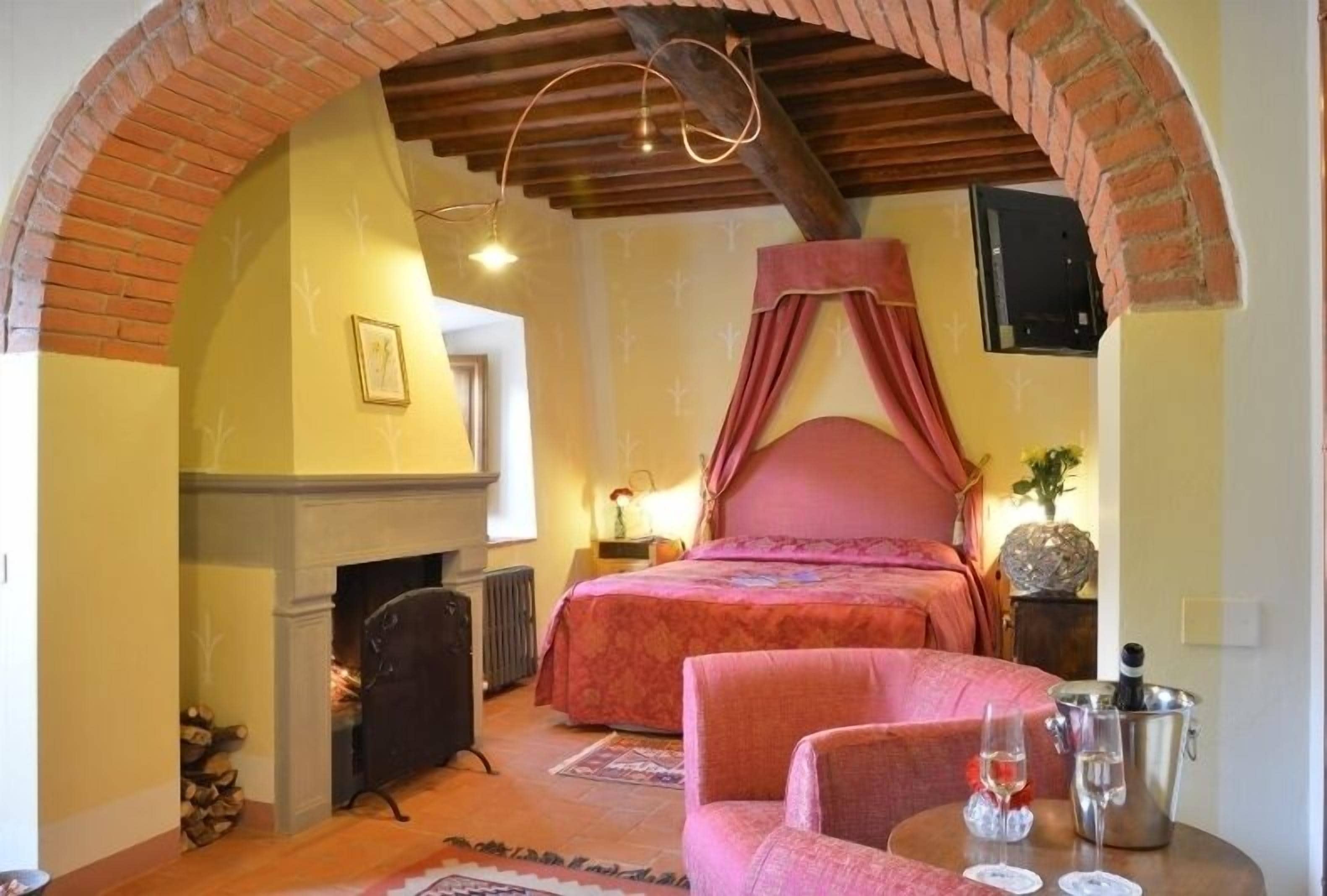 A bedroom with a brick archway, a fireplace, a bed with a canopy, a chair, and a table with glasses and a bottle.