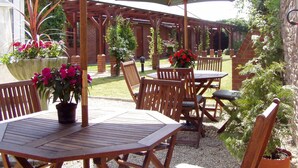 Un patio al aire libre con mesas y sillas de madera, rodeado de muros de ladrillo y plantas en macetas.