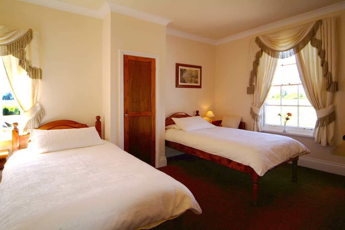 Une chambre avec deux lits simples, une porte en bois, un tableau encadré au mur et une fenêtre avec des rideaux.