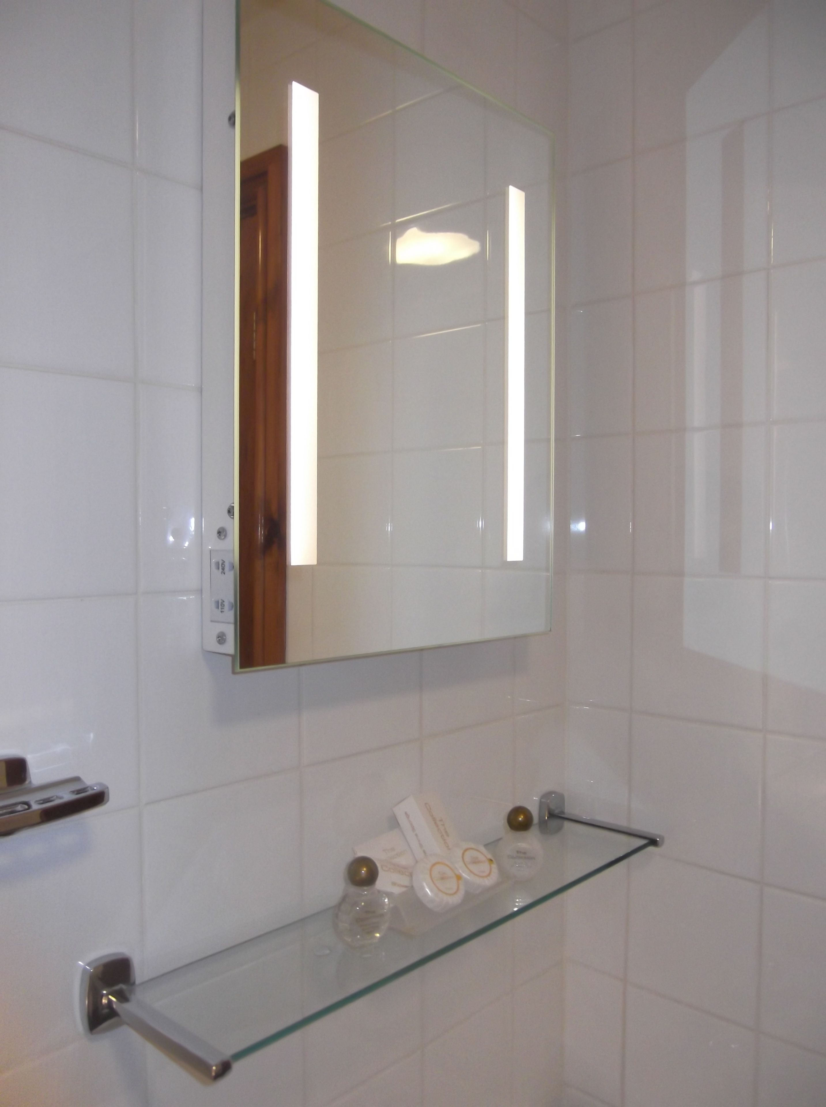 A bathroom with white tiles, a glass shelf with toiletries, and a mirror reflecting a door.