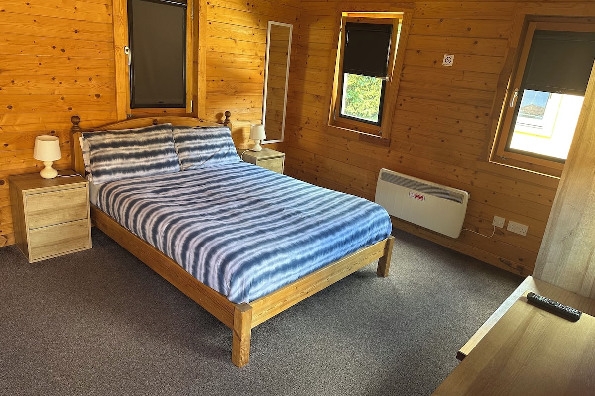 A wooden cabin room with a bed, two bedside tables, a window, and a radiator.