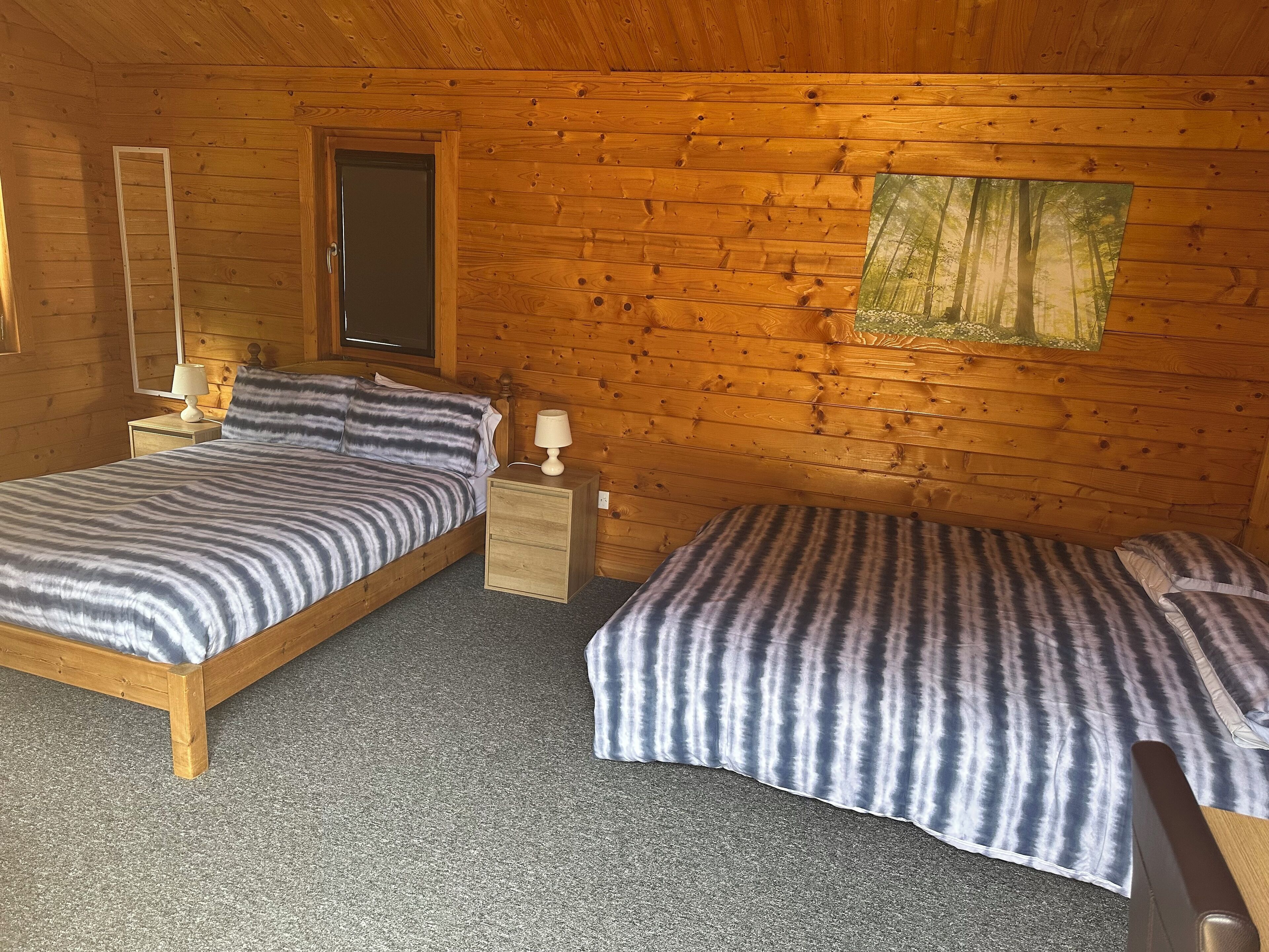 A wooden cabin room with two beds, a nightstand, a mirror, and a window with a view of trees.