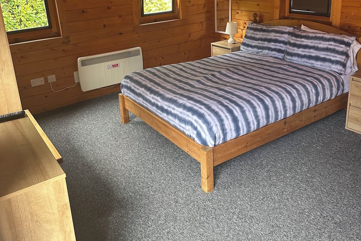 A wooden cabin bedroom with a bed, two windows, a heater, and a nightstand.
