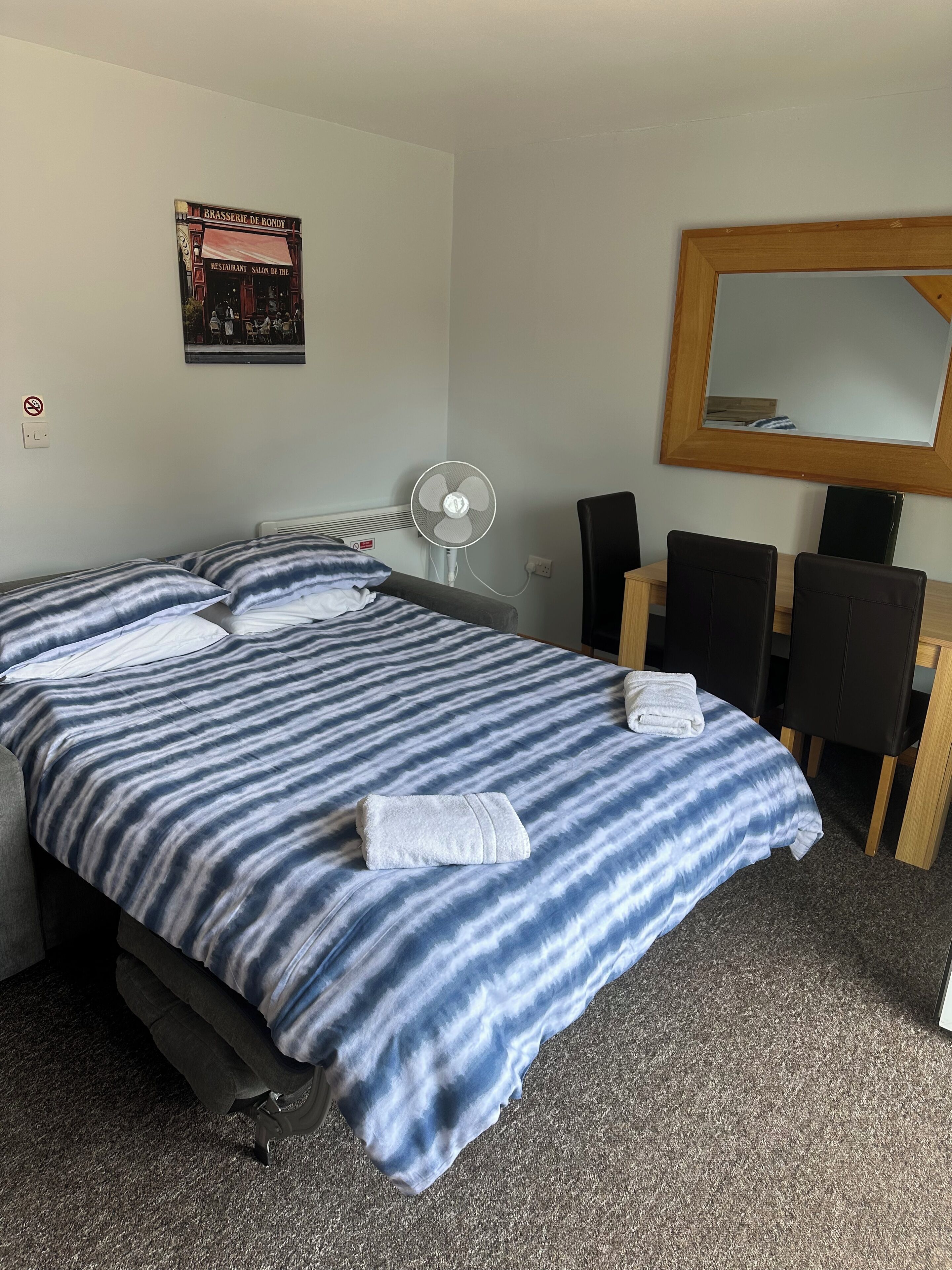 Et hotelværelse med seng, skrivebord, stol, ventilator og spejl.