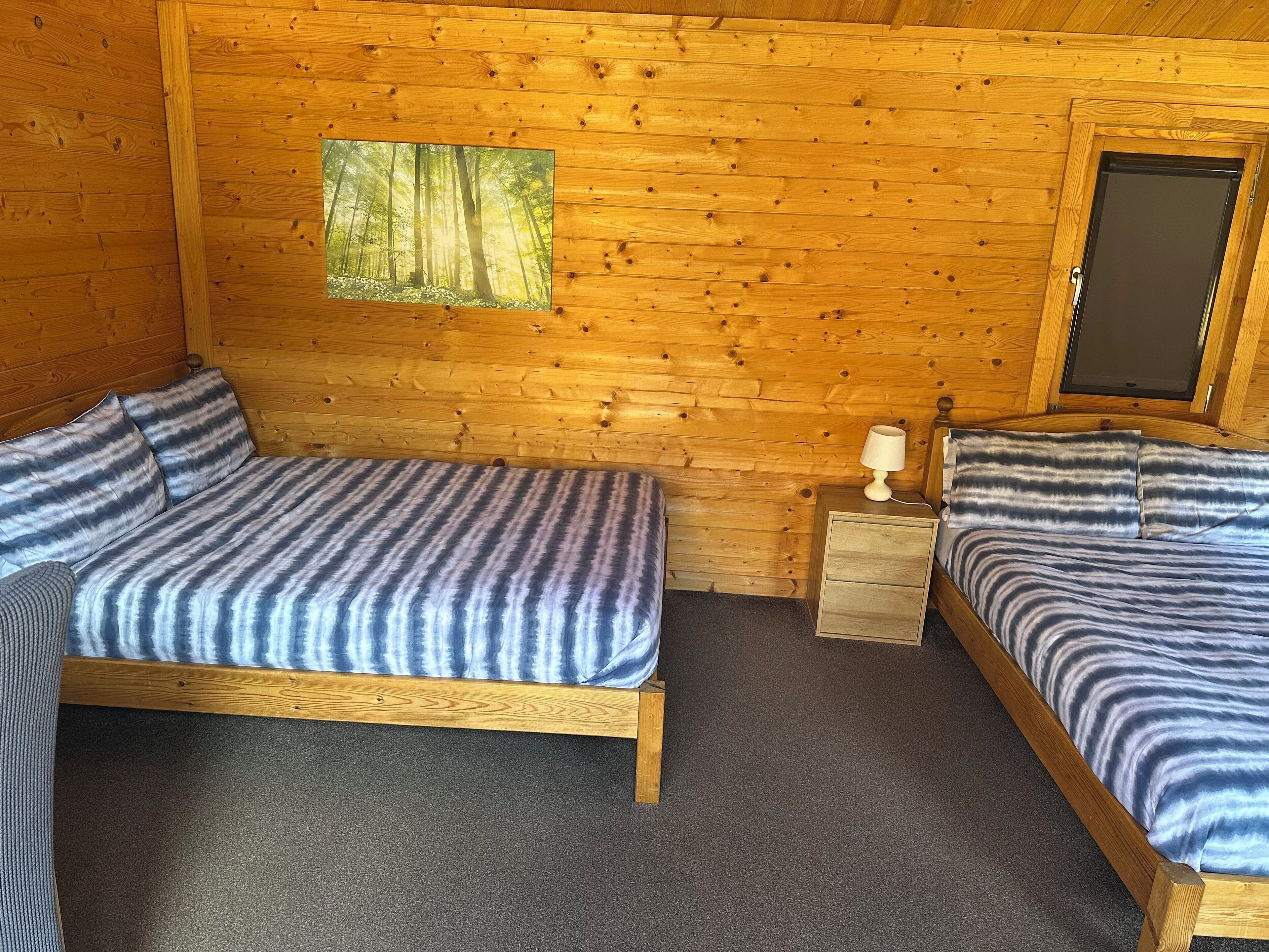 A wooden cabin room with two beds, a nightstand, a lamp, and a window with a forest view.