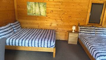 A wooden cabin room with two beds, a nightstand, a lamp, and a window with a forest view.