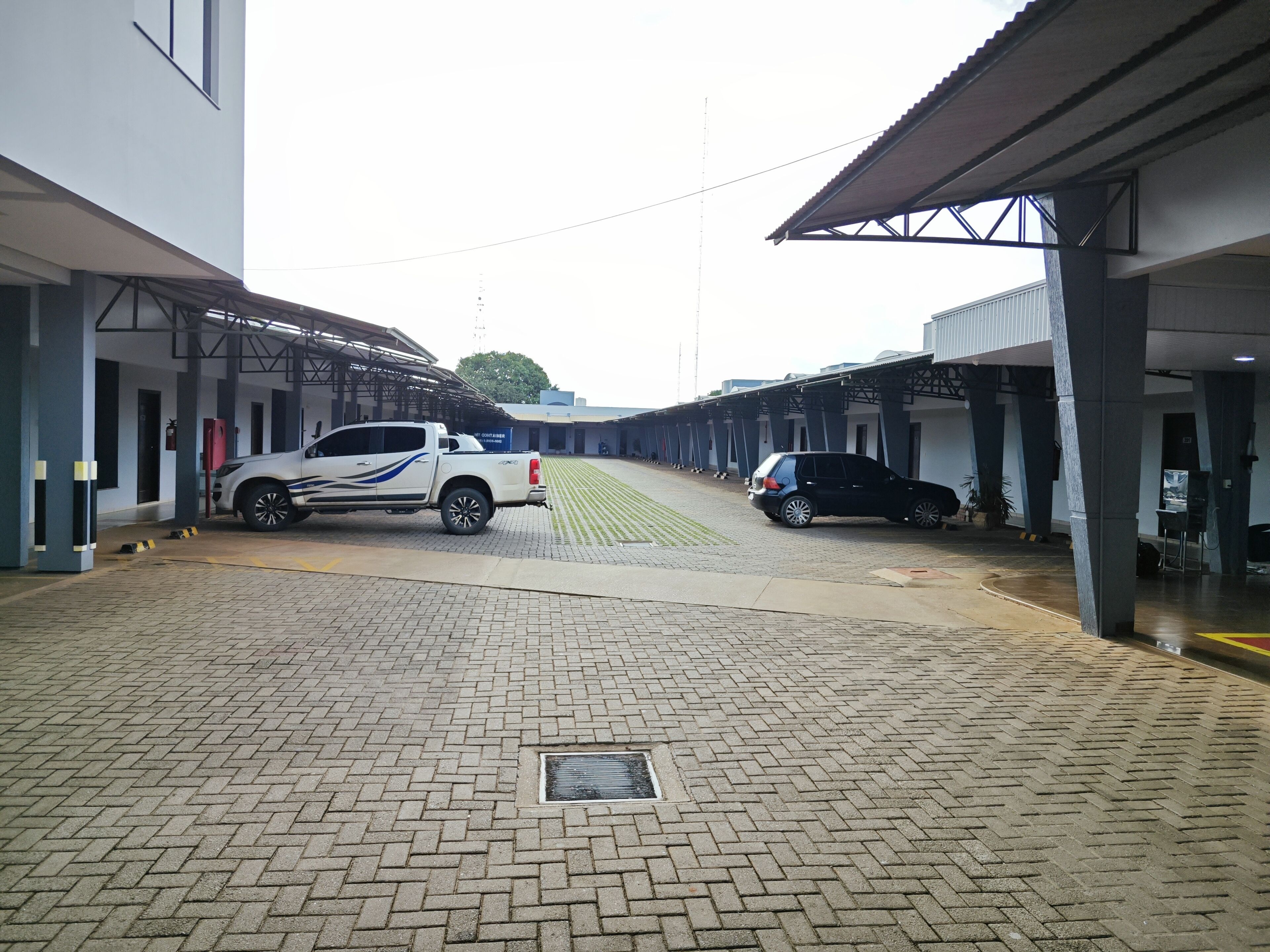 Un área de estacionamiento techada con dos vehículos aparcados, un edificio de techo plano y cielo despejado.