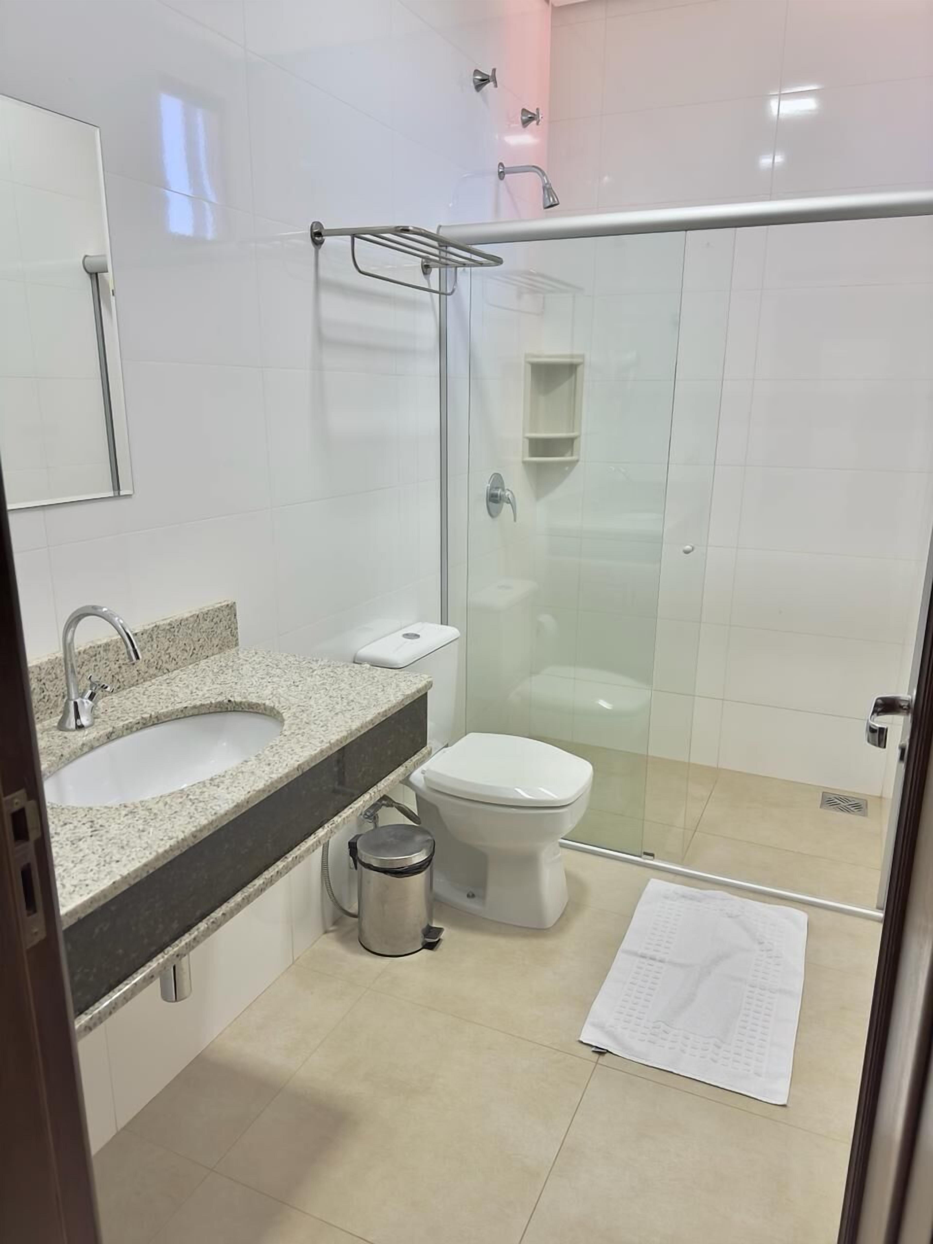 A modern bathroom with a glass-enclosed shower, a sink with a granite countertop, a toilet, and a trash can.