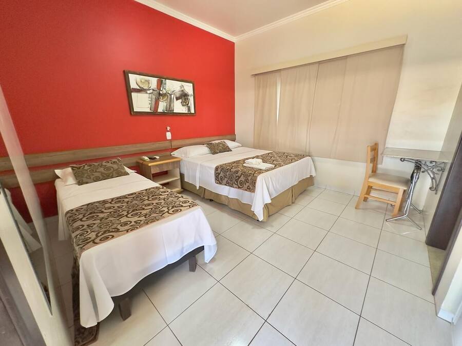 A hotel room with two beds, a red wall, a large window with curtains, and a wooden chair.