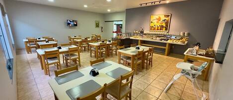 Un comedor con varias mesas y sillas, un televisor montado en la pared y una barra con diversos objetos.
