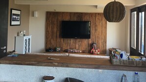 A modern living room with a large TV mounted on a wooden wall, a kitchen area with a sink and countertop, and a dining area with a bench.