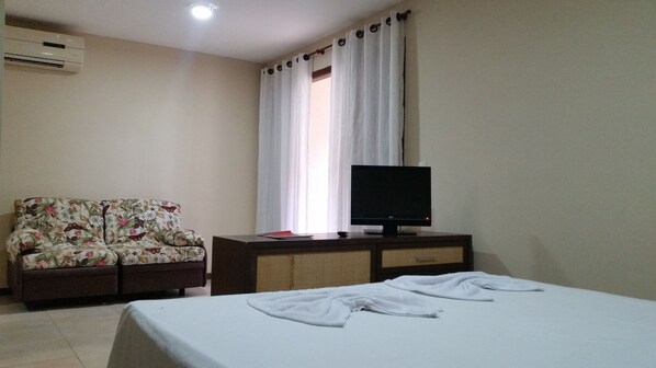 A bedroom with a bed, a TV on a wooden cabinet, a floral sofa, and an air conditioner on the wall. - Solar Pipa Apartamentos (Tibau do Sul)