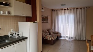 A kitchen with white cabinets, a refrigerator, and a dining area with a sofa and curtains. - Solar Pipa Apartamentos (Tibau do Sul)