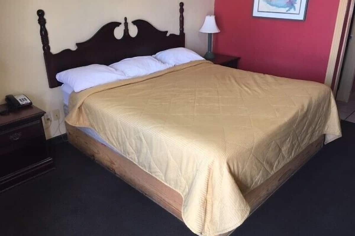 Uma cama de solteiro com cabeceira de madeira, um cobreleito bege e uma mesinha de cabeceira com abajur e telefone.
