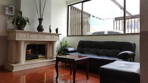 A living room with a fireplace, a black leather sofa, a wooden coffee table, and a large window with blinds. - Hotel Mont Bleu (Bogotá)